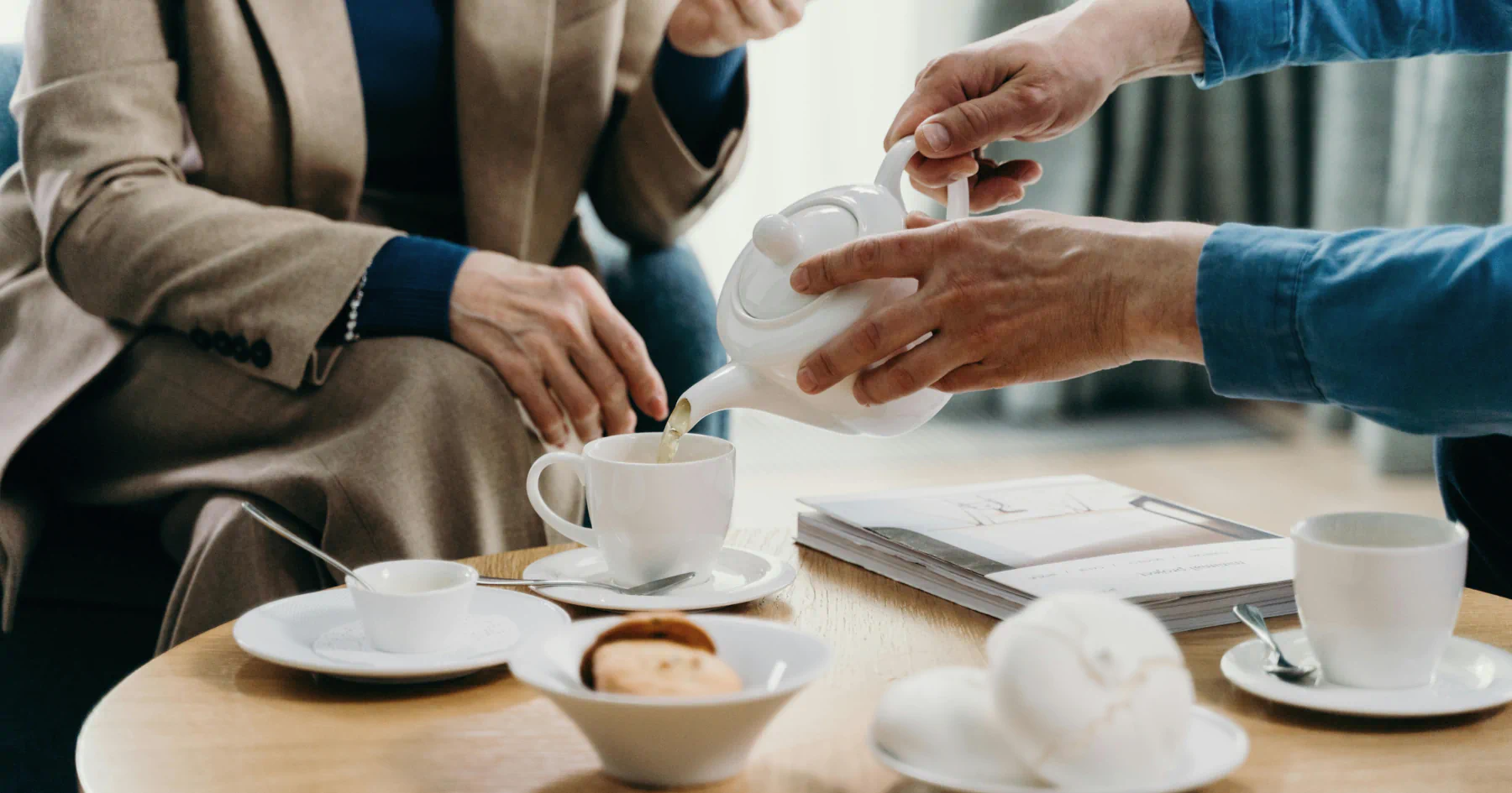 Persoon schenkt thee in een kopje tijdens een zakelijke meeting met koffie en koekjes.
